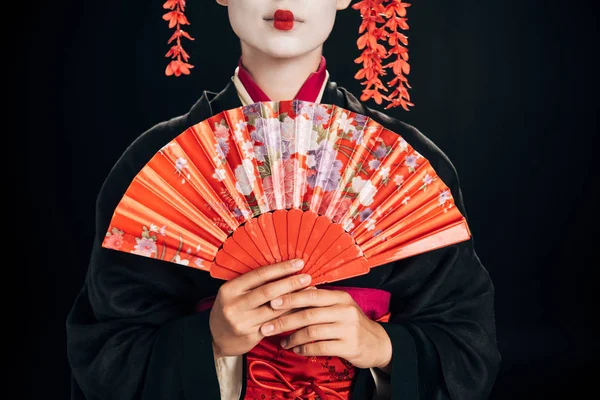 Vista ritagliata di bella geisha in kimono nero con fiori rossi in capelli tenendo tradizionale ventilatore a mano isolato su nero — Foto stock