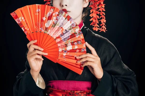 Частковий вид на красиву гейшу в чорному кімоно з червоними квітами у волоссі, що тримає традиційний вентилятор рук ізольовано на чорному — стокове фото