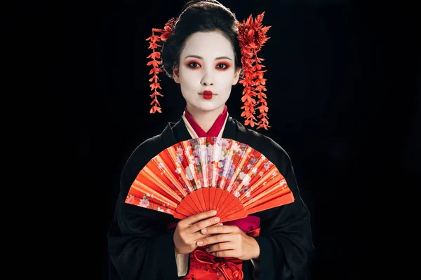 Bela gueixa em quimono preto com flores vermelhas no cabelo segurando fã mão tradicional isolado em preto — Fotografia de Stock