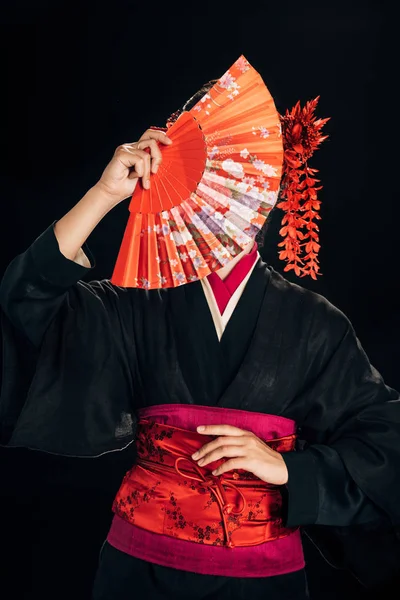 Wunderschöne Geisha im schwarzen Kimono mit roten Blumen im Haar, die den traditionellen Handfächer vor das Gesicht hält, isoliert auf schwarz — Stockfoto