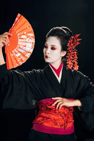 Belle geisha en kimono noir avec des fleurs rouges dans les cheveux tenant ventilateur main traditionnelle isolé sur noir — Photo de stock