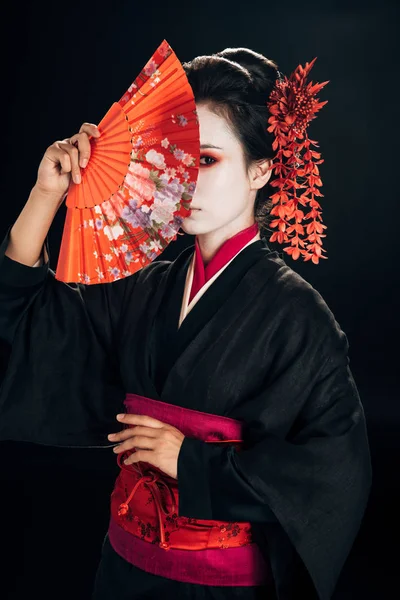 Schöne Geisha im schwarzen Kimono mit roten Blumen im Haar versteckt sich hinter traditionellen Handfächer isoliert auf schwarz — Stockfoto