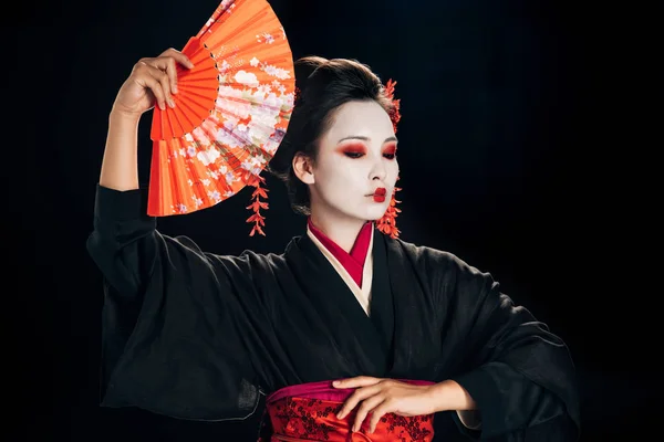 Schöne Geisha im schwarzen Kimono mit roten Blumen im Haar, die den traditionellen orangefarbenen Handfächer isoliert auf schwarz hält — Stockfoto