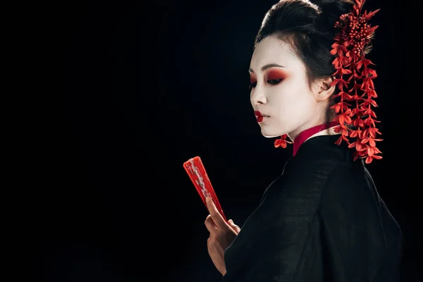 Wunderschöne Geisha im schwarzen Kimono mit roten Blumen im Haar, die einen auf schwarz isolierten Handfächer hält — Stockfoto