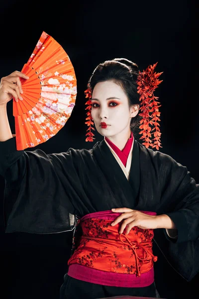 Beautiful geisha in black kimono with red flowers in hair posing with traditional hand fan isolated on black — Stock Photo