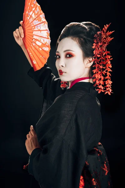 Bella geisha in kimono nero con fiori rossi in capelli gesticolando con tradizionale ventaglio a mano isolato su nero — Foto stock