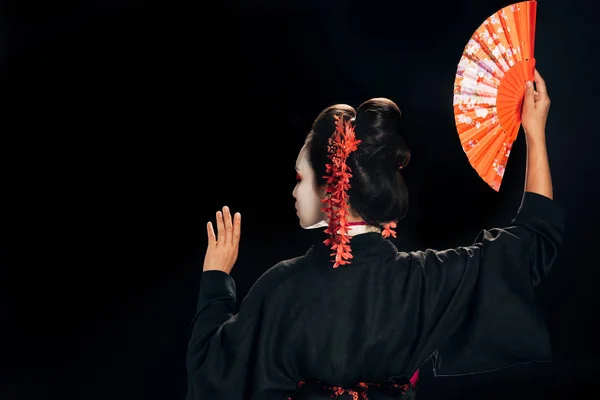 Vista posterior de hermosa geisha en kimono negro con flores rojas en el pelo sosteniendo ventilador de mano tradicional y gesto aislado en negro - foto de stock