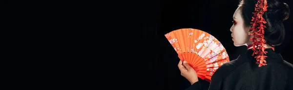 Visão traseira da bela gueixa em quimono preto com flores vermelhas no cabelo segurando ventilador tradicional mão isolado em preto, tiro panorâmico — Fotografia de Stock