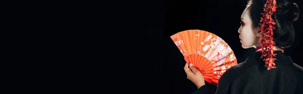 Vue arrière de la belle geisha en kimono noir avec des fleurs rouges dans les cheveux tenant ventilateur traditionnel isolé sur noir, panoramique — Photo de stock