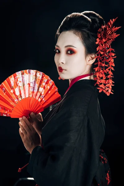 Gueixa em quimono preto com flores vermelhas no cabelo segurando ventilador de mão tradicional isolado em preto — Fotografia de Stock