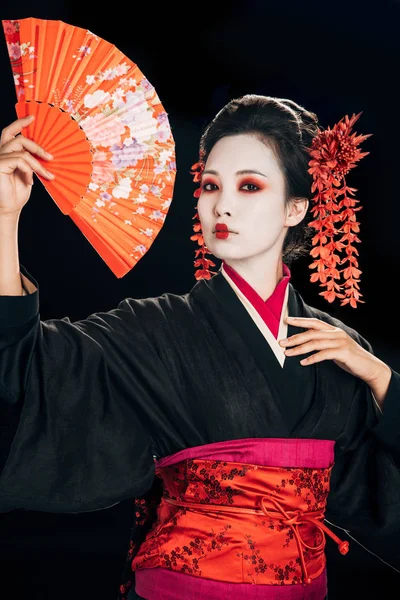 Hermosa geisha en kimono negro con flores rojas en el pelo sosteniendo ventilador de mano tradicional aislado en negro - foto de stock