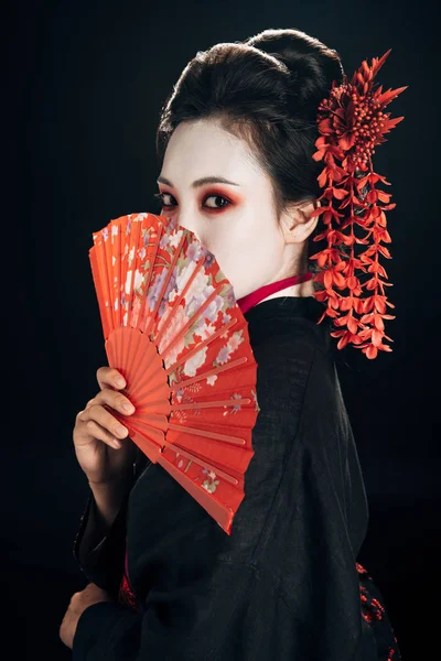 Красива гейша в чорному кімоно з червоними квітами у волоссі, що тримає традиційний вентилятор рук ізольовано на чорному — стокове фото