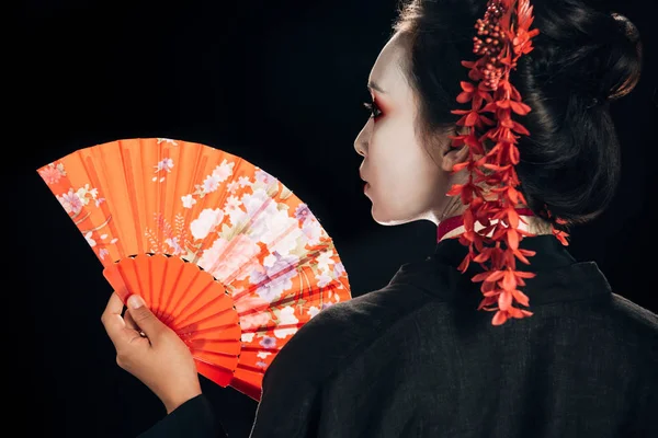 Vue arrière de la belle geisha en kimono noir avec des fleurs rouges dans les cheveux tenant ventilateur traditionnel isolé sur noir — Photo de stock