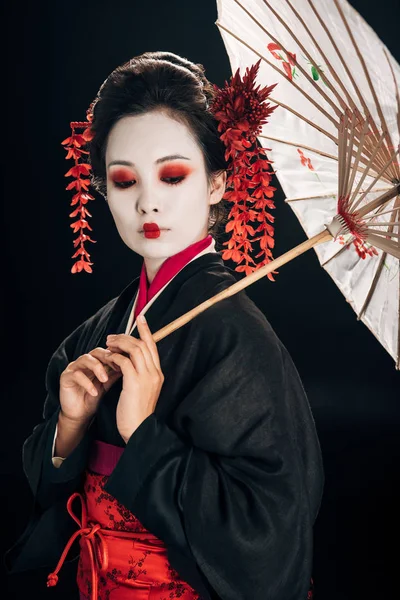 Красивая гейша в черном кимоно с красными цветами в волосах, держа традиционный азиатский зонтик изолирован на черном — стоковое фото