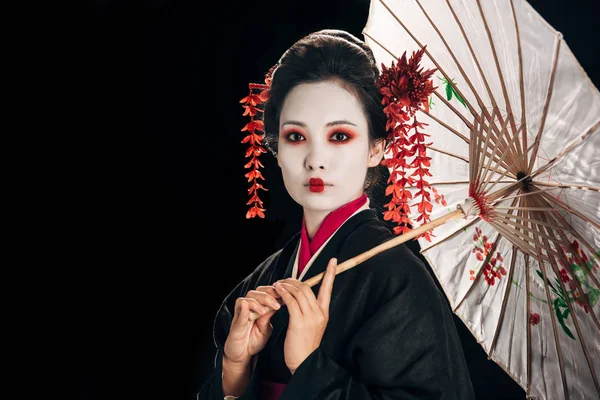Hermosa geisha en kimono negro con flores rojas en el pelo sosteniendo paraguas asiático tradicional aislado en negro con espacio de copia - foto de stock