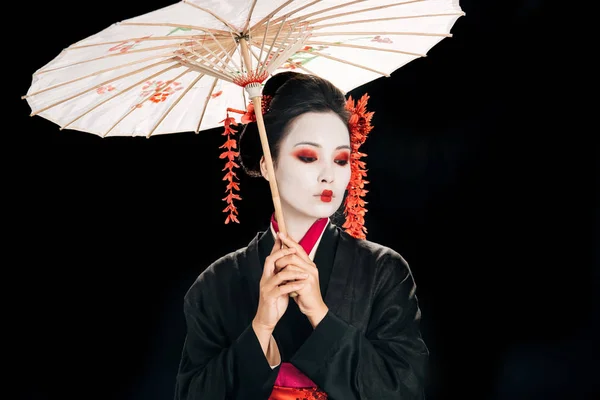 Красивая гейша в черном кимоно с красными цветами в волосах держа традиционный азиатский зонтик и глядя вниз изолированы на черный — стоковое фото