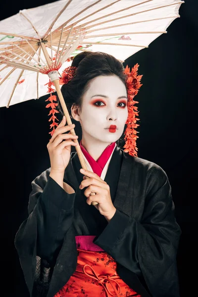 Hermosa geisha en kimono con flores rojas en el pelo sosteniendo paraguas asiático tradicional aislado en negro - foto de stock