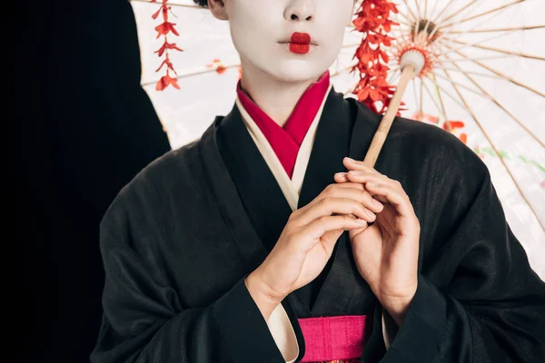 Частковий вид на красиву гейшу в чорному кімоно з червоними квітами у волоссі, що тримає традиційну азіатську парасольку ізольовано на чорному — стокове фото