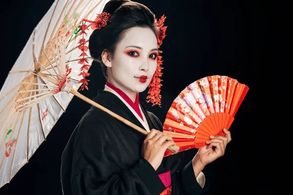 Bela gueixa no preto quimono com flores vermelhas no cabelo segurando asiático guarda-chuva e mão fã isolado no preto — Fotografia de Stock