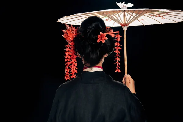 Vue arrière de la belle geisha en kimono noir avec des fleurs rouges dans les cheveux tenant parapluie asiatique traditionnel isolé sur noir — Photo de stock