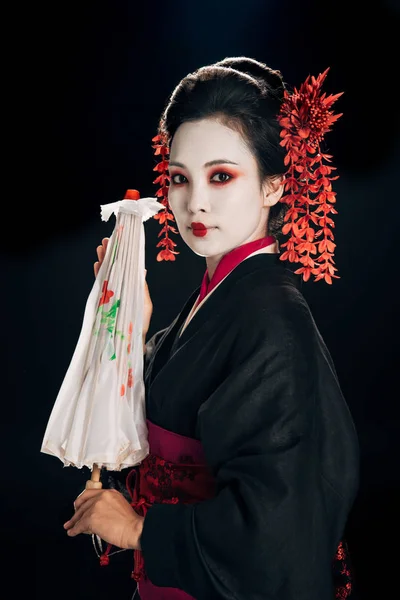 Belle geisha en kimono noir avec des fleurs rouges dans les cheveux tenant parapluie asiatique traditionnel isolé sur noir — Photo de stock
