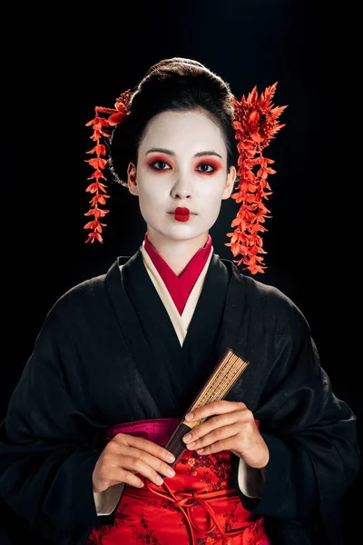 Hermosa geisha en kimono negro y rojo y flores en el pelo sosteniendo ventilador de mano de madera aislado en negro - foto de stock