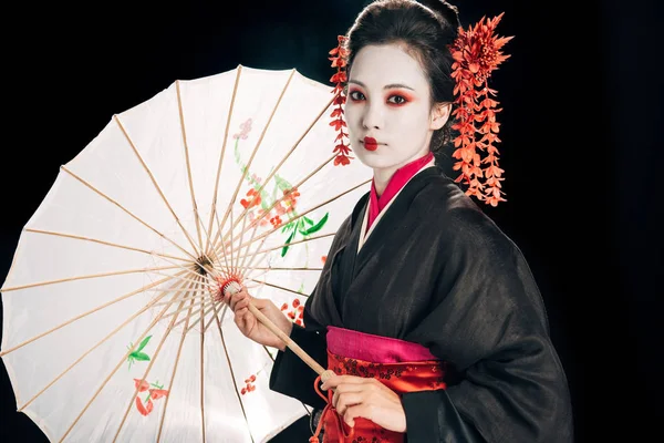 Geisha en kimono negro con flores rojas en el pelo sosteniendo paraguas asiático tradicional aislado en negro - foto de stock