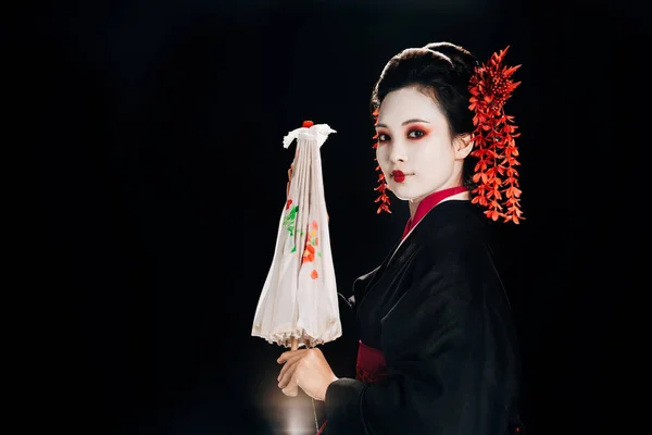 Боковой вид гейши в черном кимоно с красными цветами в волосах, держащих традиционный азиатский зонтик изолирован на черном — стоковое фото