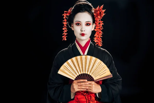 Geisha in schwarzem Kimono mit roten Blumen im Haar hält traditionellen asiatischen Handfächer isoliert auf schwarz — Stockfoto