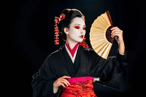 Geisha in kimono with red flowers in hair holding traditional asian hand fan isolated on black — Stock Photo