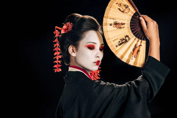 Vue latérale de la geisha en kimono noir avec des fleurs rouges dans les cheveux tenant traditionnel asiatique ventilateur à main isolé sur noir — Photo de stock