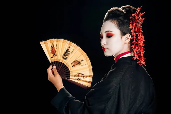 Geisha im schwarzen Kimono mit roten Blüten im Haar schaut auf den traditionellen asiatischen Handfächer isoliert auf schwarz — Stockfoto