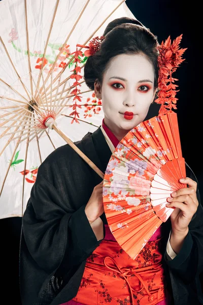 Sonriente hermosa geisha en kimono negro con flores rojas en el pelo sosteniendo paraguas asiático tradicional y ventilador de mano aislado en negro - foto de stock