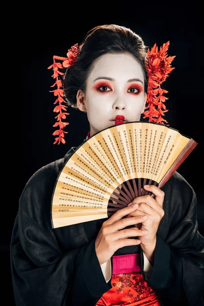 Jovem gueixa no preto quimono com flores vermelhas no cabelo segurando tradicional asiático mão fã isolado no preto — Fotografia de Stock