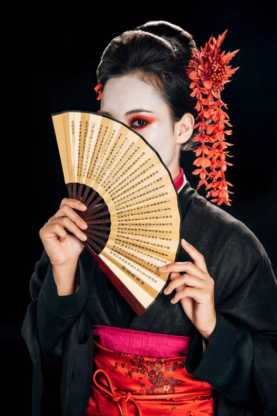 Geisha en kimono noir avec des fleurs rouges dans les cheveux et le visage obscur tenant ventilateur main asiatique traditionnelle isolé sur noir — Photo de stock