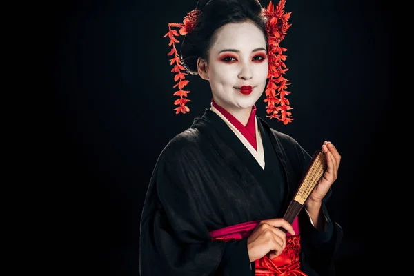 Heureux geisha en kimono noir avec des fleurs rouges dans les cheveux tenant traditionnelle asiatique main ventilateur isolé sur noir — Photo de stock