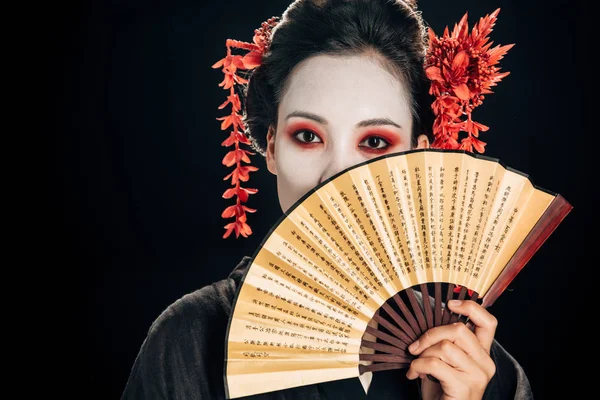 Гейша в черном кимоно с красными цветами в волосах держа традиционный азиатский вентилятор руки возле лица изолированы на черном — стоковое фото