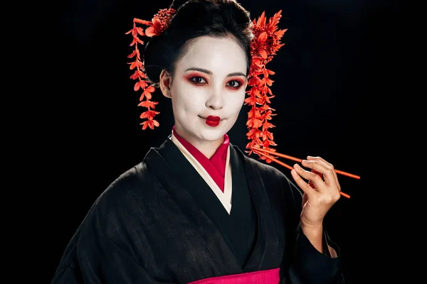 Belle geisha souriante en kimono noir avec des fleurs rouges dans les cheveux tenant des baguettes isolées sur noir — Photo de stock