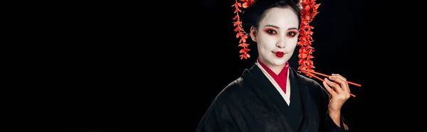 Улыбающаяся красивая гейша в черном кимоно с красными цветами в волосах, держащая палочки, изолированные на черном, панорамном снимке — стоковое фото