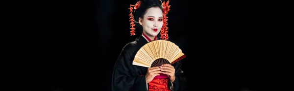 Улыбающаяся гейша в черном кимоно с красными цветами в волосах, держащая традиционный ручной вентилятор, изолированный на черном, панорамный снимок — стоковое фото