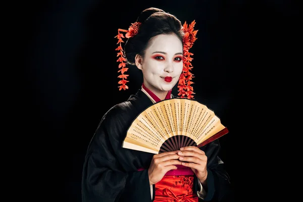 Fröhliche Geisha im schwarzen Kimono mit roten Blumen im Haar, die einen traditionellen asiatischen Handfächer isoliert auf schwarz hält — Stockfoto