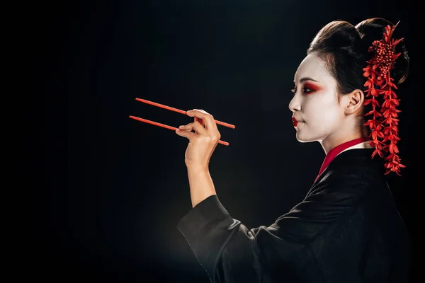 Vue latérale de la belle geisha en kimono noir avec des fleurs rouges dans les cheveux tenant des baguettes isolées sur noir — Photo de stock