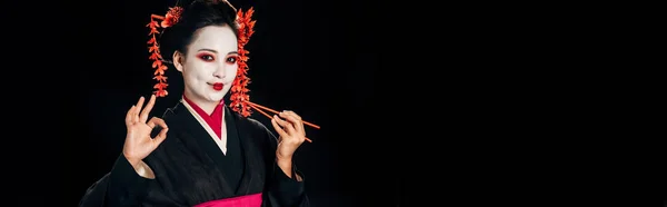Belle geisha souriante en kimono noir avec des fleurs rouges dans les cheveux tenant des baguettes et montrant OK signe isolé sur noir, plan panoramique — Photo de stock