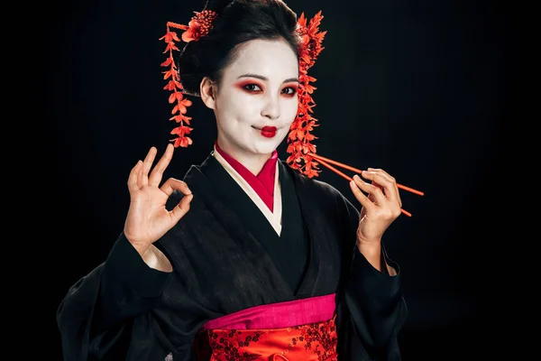 Smiling beautiful geisha in black kimono with red flowers in hair holding chopsticks and showing okay sign isolated on black — Stock Photo
