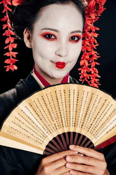 Портрет улыбающейся красивой гейши в черно-красном кимоно и цветы в волосах, держащих ручной вентилятор, изолированный на черном — стоковое фото