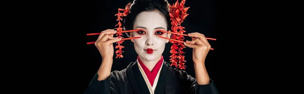 Hermosa geisha en kimono negro con flores rojas en el pelo sosteniendo palillos cerca de los ojos aislados en negro, plano panorámico - foto de stock