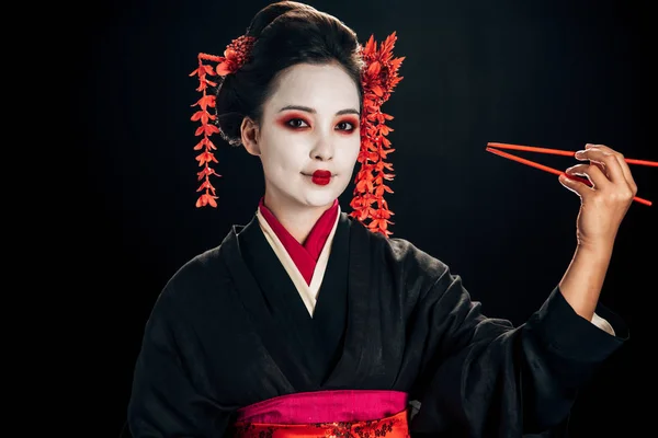 Cheerful beautiful geisha in black kimono with red flowers in hair holding chopsticks isolated on black — Stock Photo