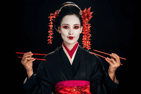 Heureuse belle geisha en kimono noir avec des fleurs rouges dans les cheveux tenant des baguettes isolées sur noir — Photo de stock