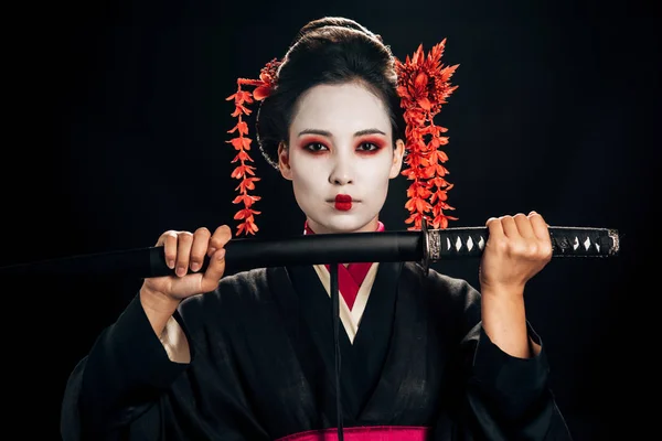Belle geisha en kimono noir avec des fleurs rouges dans les cheveux tenant katana isolé sur noir — Photo de stock