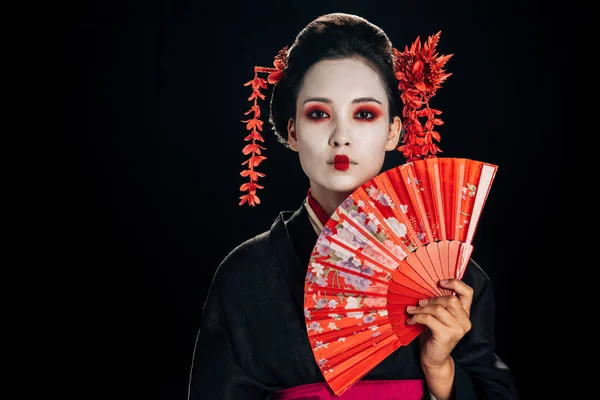 Attraktive Geisha im schwarzen Kimono mit Blumen im Haar hält helle Hand Fächer isoliert auf schwarz mit Kopierraum — Stockfoto